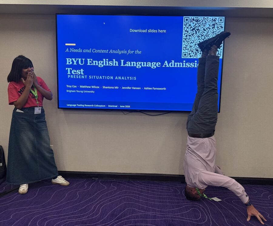 Troy Cox doing a handstand beside a presentation slide at the Language Testing Research Colloquium in Montreal, as a colleague laughs